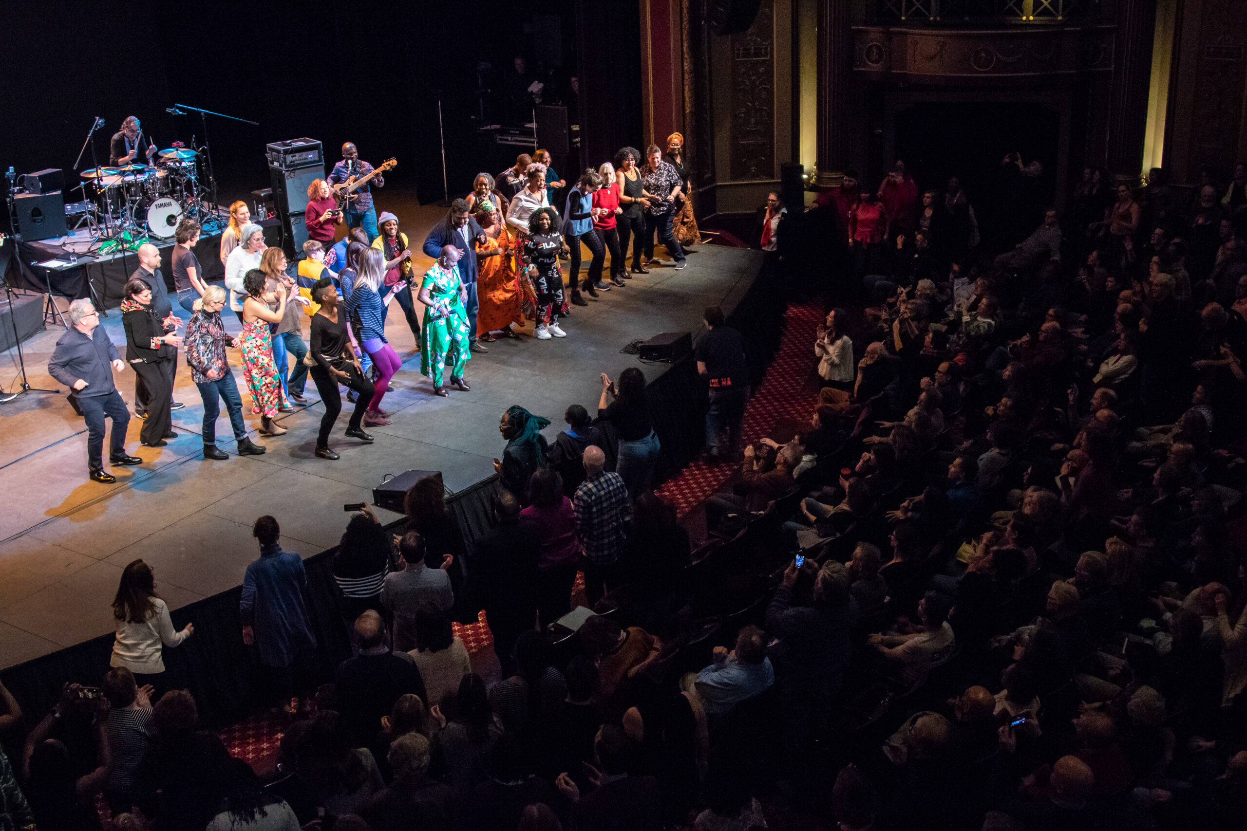 Audience members dance with Angélique Kidjo on stage at The VETS in February 2020. The first seven rows of audience members are shown dancing along.
