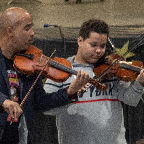A Black man with a violin instructing a young man with a violin
