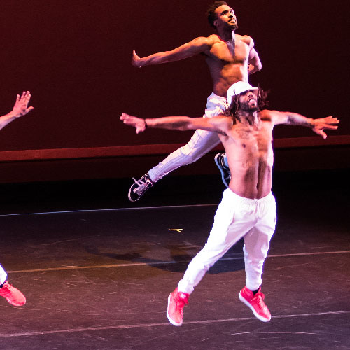 Three Black male dancers in white pants and red sneakers leaping high on a stage with their arms outstretched
