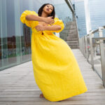 A Black woman in a full length yellow dress with puff sleeves dancing outside on a wooden boardwalk next to a glass building