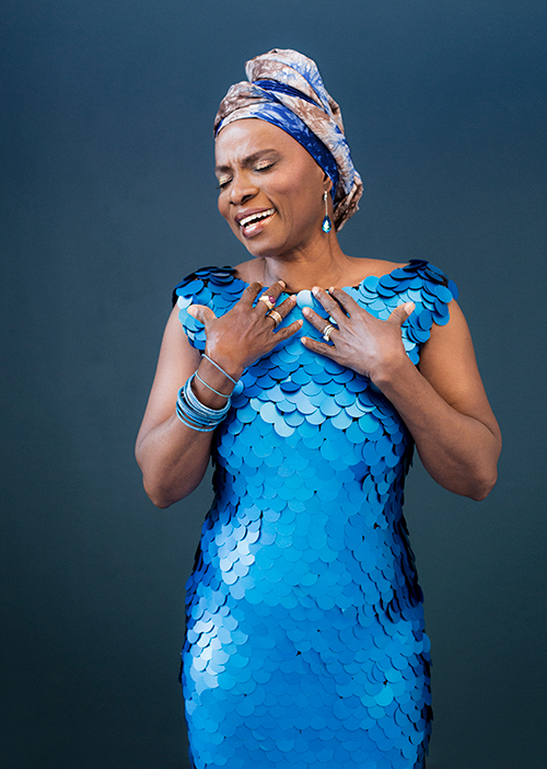 Una mujer negra con un vestido azul brillante de escamas de sirena canta con los ojos cerrados