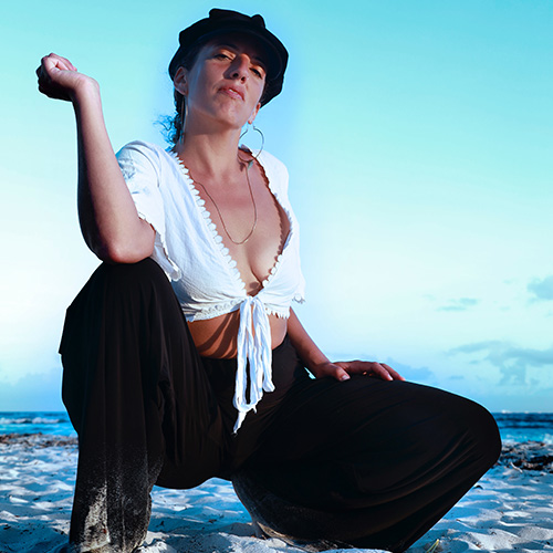 A woman in a low cut white blouse, black hat and harem pants strikes a power pose on a sunny beach