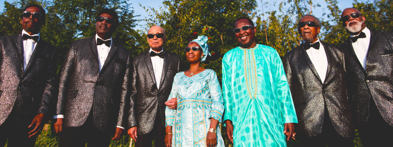Five men in black tuxedos and bowties flanking a man and a woman wearing turquoise West African robes. All are standing outdoors on a sunny day and wearing dark glasses