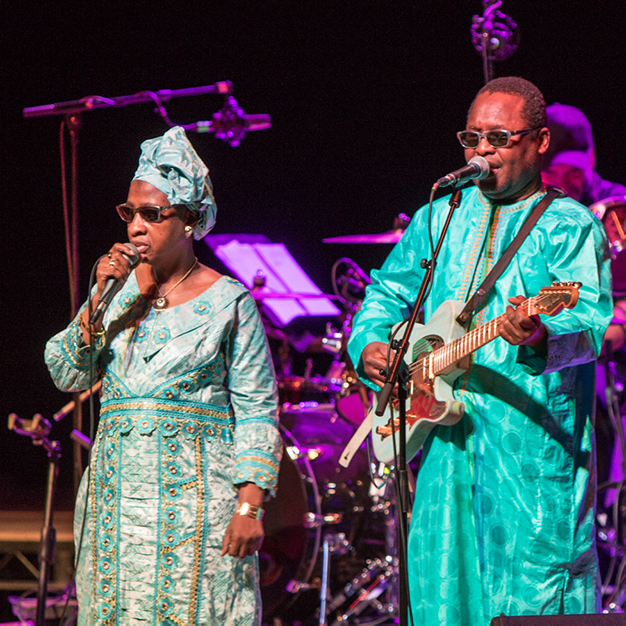 A Black woman and a Black man singing on a stage wearing dark glasses and turquoise robes