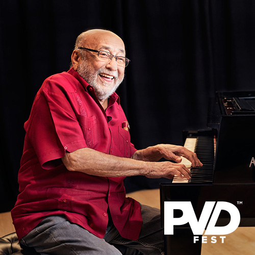 A man with a silver beard, red short sleeve collared shirt and glasses smiling while playing a black piano