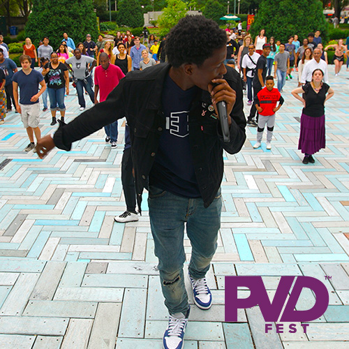 A young Black man holding a mic demonstrating some dance moves to a crowd of people on an outdoor brick plaza