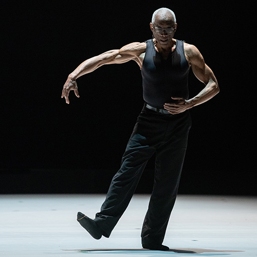 An older Black dancer in black pants and tank top poses alone on a darkened stage