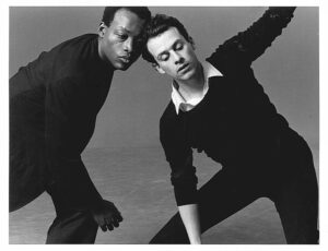 Two dancers, a Black man and a white man, pictured in black and white lean in towards each other with their heads together and their feet apart