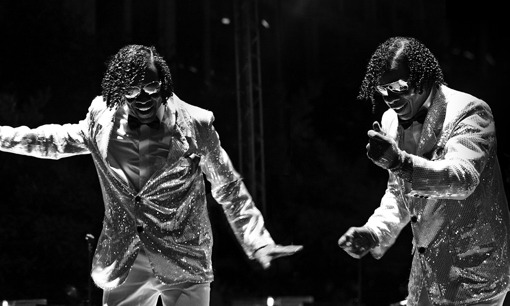 A black and white image of two identical Black adult male dancers wearing sequined suits and sunglasses dancing under some spotlights