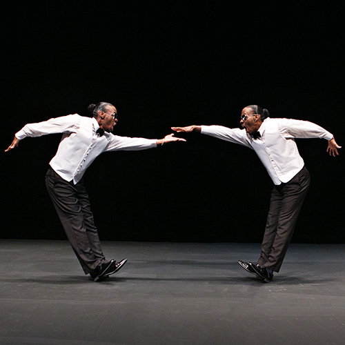 Identical twin Black adult men wearing white button down shirts and black pants and shoes face each other on a dark stage. They are both leaning back on their heels and each has a hand extended out to the other although not touching