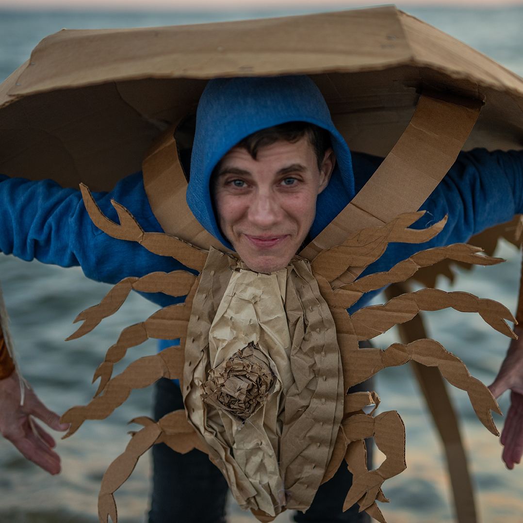 A young white man wearing a blue hoodie and an elaborate cardboard costume of a horseshoe crab stands facing away from the sea with his arms in a crab stance