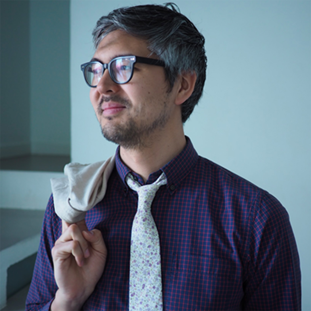 A middle-aged Asian man with salt and pepper hair and light goatee smiles in three-quarter profile. He wears a blue and red checked shirt, a white floral tie and glasses