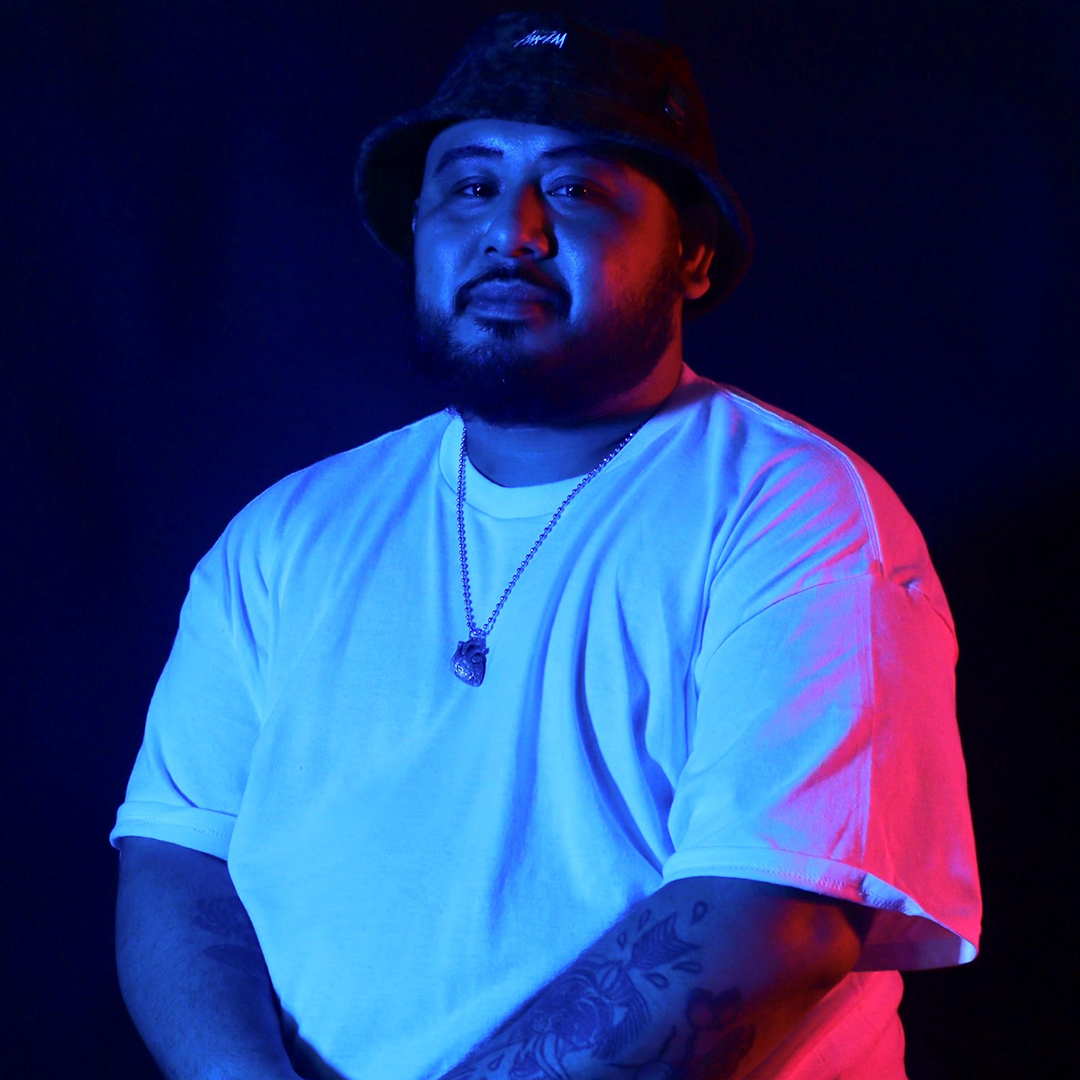 A heavyset Cambodian man wearing a black brimmed hat, white t-shirt and silver necklace stands with his arms lightly crossed against a dark background lit with blue and red theatrical lights