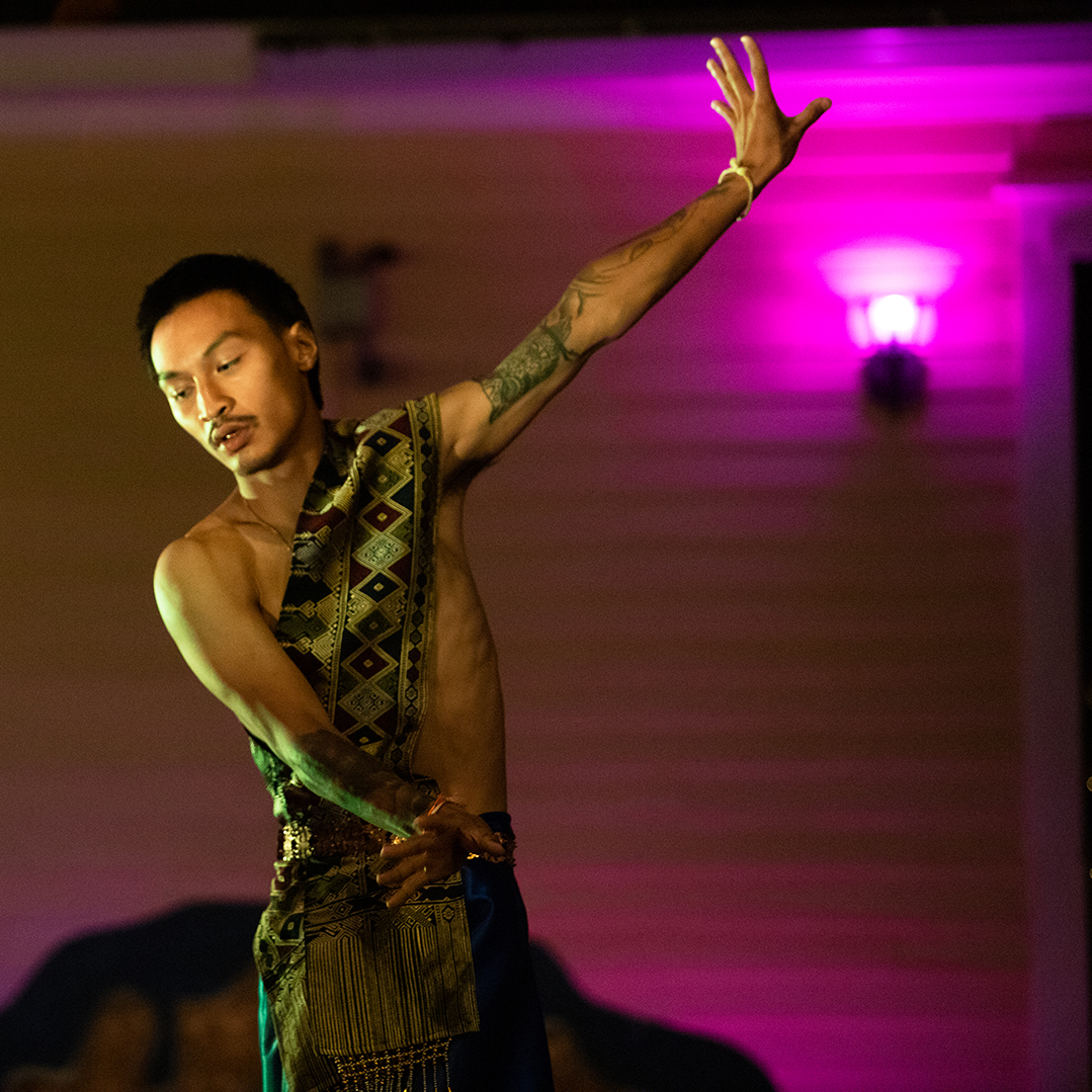 A young Asian man dancing shirtless outside in front of a yellow sided house with a pink outdoor light