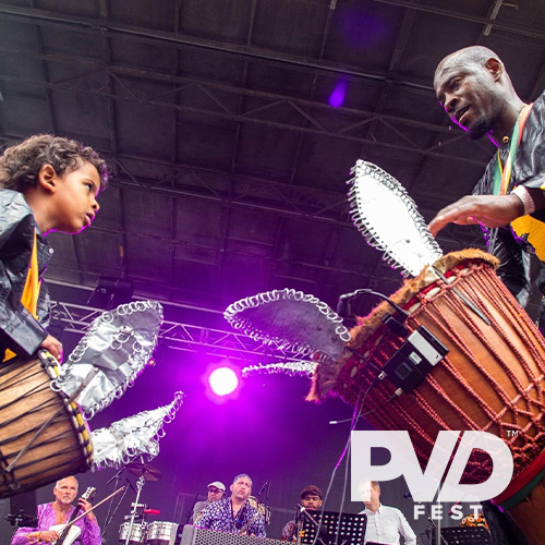 The artist Sidy Maiga plays a wooden djembe handdrum facing his young son who is playing a smaller version of the drum. PVDFest logo overlaid in white.