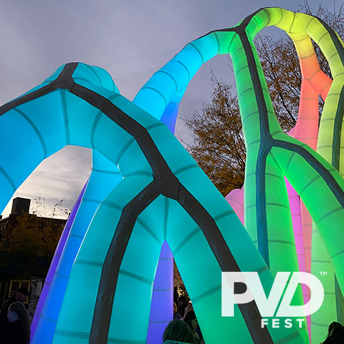 A large inflated sculpture of intersecting arches lit up in rainbow colors in an outdoor location at dusk. PVDFest logo overlaid in white.