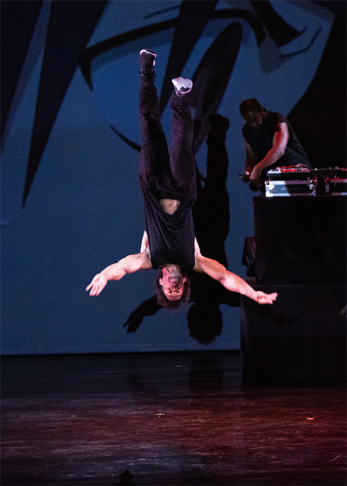 A young male dancer doing a flip in midair while a DJ spins records behind him