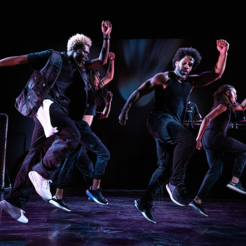 A group of Black male dancers dressed in black dancing facing the viewer in three quarter profile with their faces in shadow