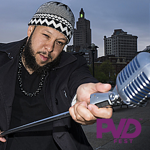 A bearded man in a black and white knit cap holds a metal clad stage mic towards the viewer he is outside with the Providence skyline behind him