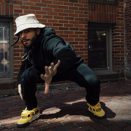 A man in a white bucket hat and hello sneakers strikes a crouching dance pose outdoors in front of a brick wall