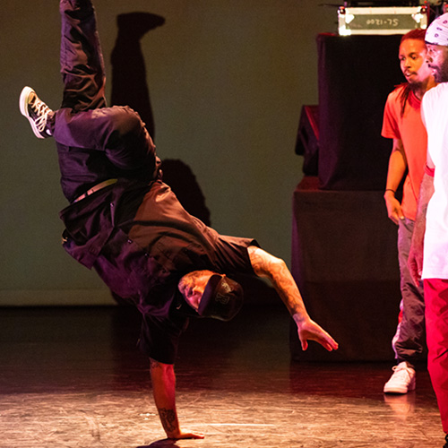 A male dancer dressed in black does a one hand handstand in a twist while men stand to either side looking on