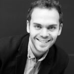 A black and white portrait of a smiling white man with short cropped hair he is wearing a collared shirt and black blazer