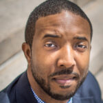 A close-up portrait of a serious looking Black male with close cropped hair and close-shaved goatee he is wearing a blue blazer