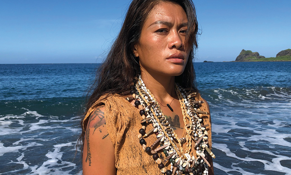 A native Taiwanese woman with long brown hair wearing a leather top and many beaded necklaces stands outside on the shore of a foamy ocean on a sunny day