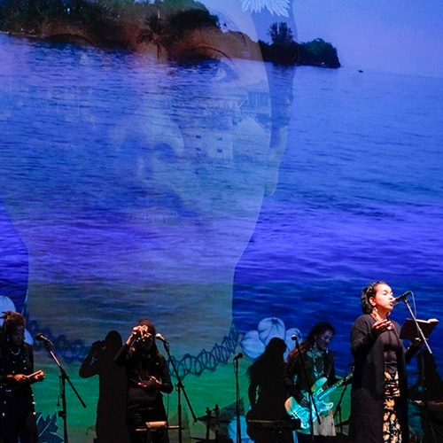 A movie screen with an image of the sea with a large face superimposed over it is the backdrop for a live band of musicians performing