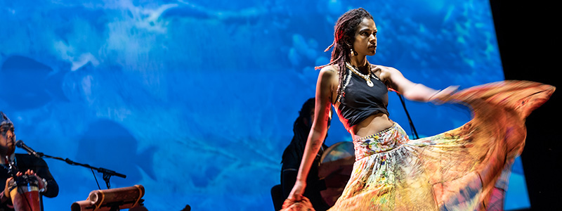 A woman in a flowy yellow skirt and grey halter top dances in front of musicians with a backdrop of underwater film footage behind them