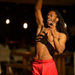 A shirtless young Black man with long hair singing with his left arm raised high into the air