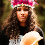 A young Indigenous woman with long curly brown hair, a tattoo on her chin wearing a pink feathered headdress and holding a large cream colored conch shell