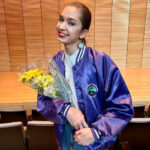 A young Indigenous woman with light brown hair pulled back in a low ponytail. She is smiling and holding a bouquet of yellow flowers inside of a theater. She is wearing a shiny purple satin jacket.