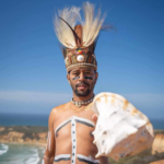 An Indigenous man wearing a headdress and body paint stands outside holding a large white conch shell