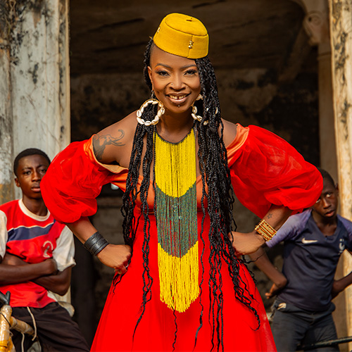 An African woman with long locs and wearing a red dress with yellow and olive green accents stands smiling with her fists on her hips in front of a group of little boys