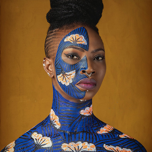 The face and shoulders of a Black woman wearing bright blue body paint with a white and ochre fan motif.