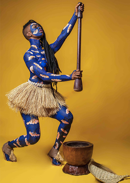 A Black woman wearing bright blue body paint with a white and ochre fan motif. She has her head thrown back and is holding a tall wooden rod or pestle above a wooden bowl that is placed on the ground next to a straw hat.