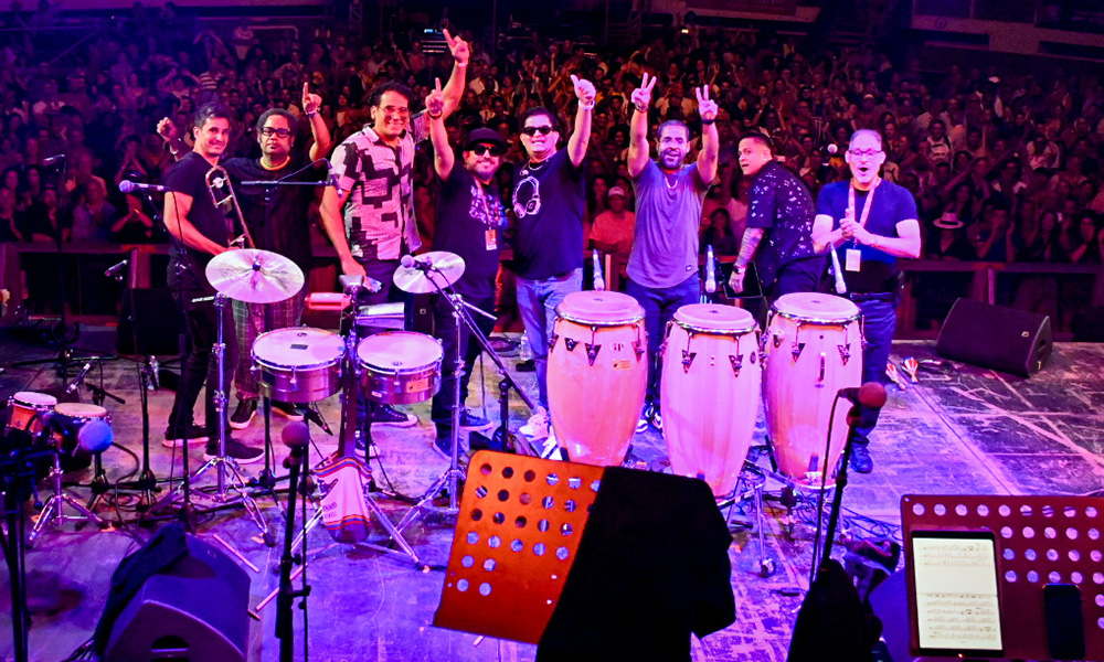 A group of eight musicians stand on a stage with their arms in the air. There are bongos in front of them and a live audience behind them.