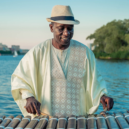 A Black musician wearing a white robe and white hat with a black band playing the ballaphone outdoors near a body of water