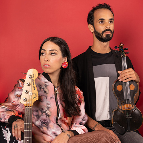 A female musician with long brown hair sits looking over her right shoulder with a bass guitar neck upright in front of her. To her left stands a tall, young male musician with short dark brown hair, beard and mustache. He is holding a dark brown violin in his left hand with light brown shapes on it that make it look like a face.