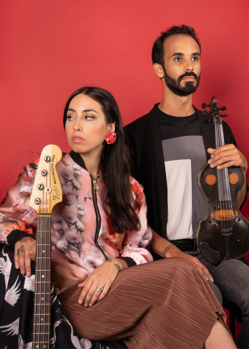 A female musician with long brown hair sits looking over her right shoulder with a bass guitar neck upright in front of her. To her left stands a tall, young male musician with short dark brown hair, beard and mustache. He is holding a dark brown violin in his left hand with light brown shapes on it that make it look like a face.