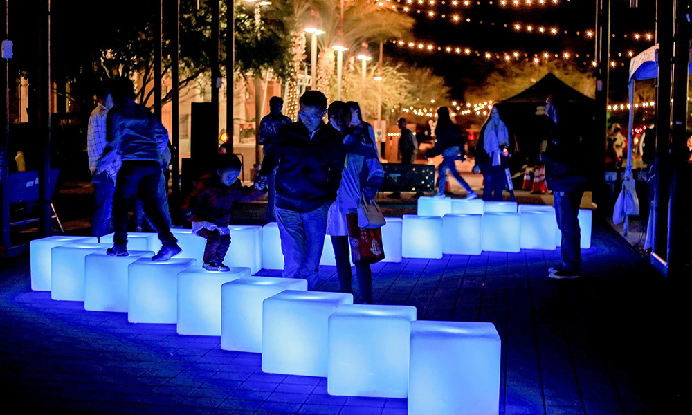 Members of the public interact with glowing blue cubes arranged in a meandering line on the ground outdoors in a park setting at night