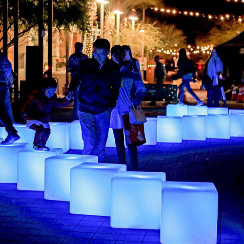 Members of the public interact with glowing blue cubes arranged in a meandering line on the ground outdoors in a park setting at night