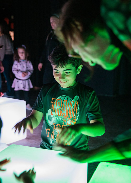 A young boy holds his hands above a glowing cube with a delighted look on his face. There are adults and other children in the foreground and background