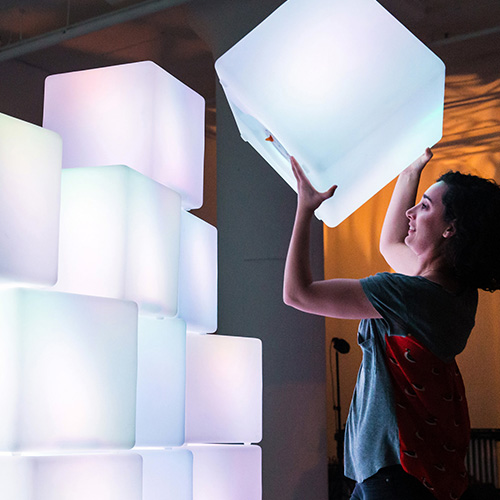 A smiling woman with curly dark hair placing a glowing white cube on a sculpture constructed from several other glowing white cubes