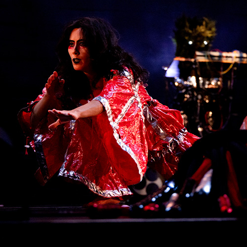 A woman in a red and white robe crouches on a dark stage reaching out towards a yellow doll suspended from a piece of fabric