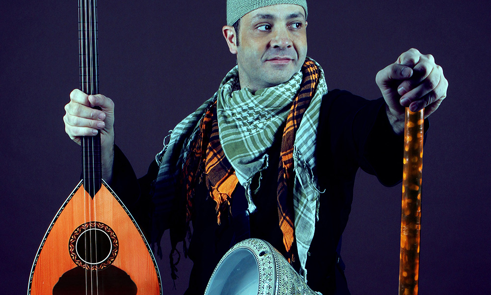 A man in a green beanie sits looking over his left shoulder. He holds in his right hand a string instrument and a staff in his left hand. A metal drum hangs from a strap around his torso.