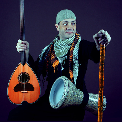 A man in a green beanie sits looking over his left shoulder. He holds in his right hand a string instrument and a staff in his left hand. A metal drum hangs from a strap around his torso.