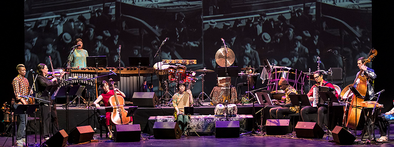 an ensemble of musicians on a stage with a black and white projection behind them.
