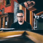A white man with salt and pepper short hair and a goatee and black glasses seated at a piano in a red wallpapered room with wooden furniture. The man's reflection is visible in the glossy black underside of the raised grand piano lid.